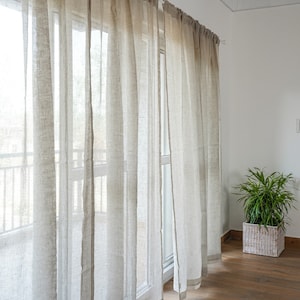 Pode incluir: Um conjunto de cortinas de linho branco translúcidas penduradas em uma janela. As cortinas são feitas de um tecido leve que permite que a luz filtre.