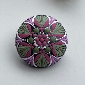 Könnte beinhalten: Ein runder, bemalter Stein mit einem Mandala-Design. Das Mandala zeigt die Farben Pink, Grün, Weiß und Schwarz.