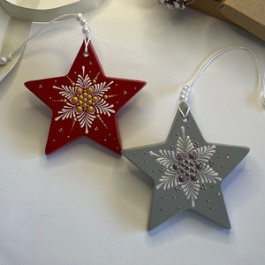May include: Two painted wooden star ornaments with white and gold snowflake designs. The red star has gold accents and the gray star has purple accents. The stars are hanging from white strings with white beads.