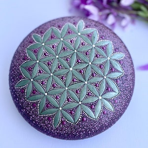 May include: A round, painted rock with a green and purple flower of life pattern. The rock is painted with small dots and has a glossy finish.