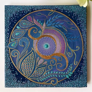 Op de afbeelding: Een cirkelvormig schilderij op een blauw canvas met een gouden, paarse en turquoise mandala-ontwerp. De mandala is omgeven door een rand van blauwe stippen.