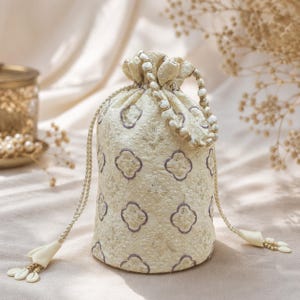 May include: A cream-coloured drawstring pouch featuring an intricate embroidered pattern of purple-outlined quatrefoil shapes. The bag includes a beaded handle and two long braided cords with decorative tassels at the ends, finished with small white beads.