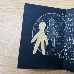 May include: A black card with a gold and blue illustration of two stick figures holding hands inside a circle. The text on the card reads "Your in dAY icl You in Per ind Sile whyy ThAT, d but if You..."