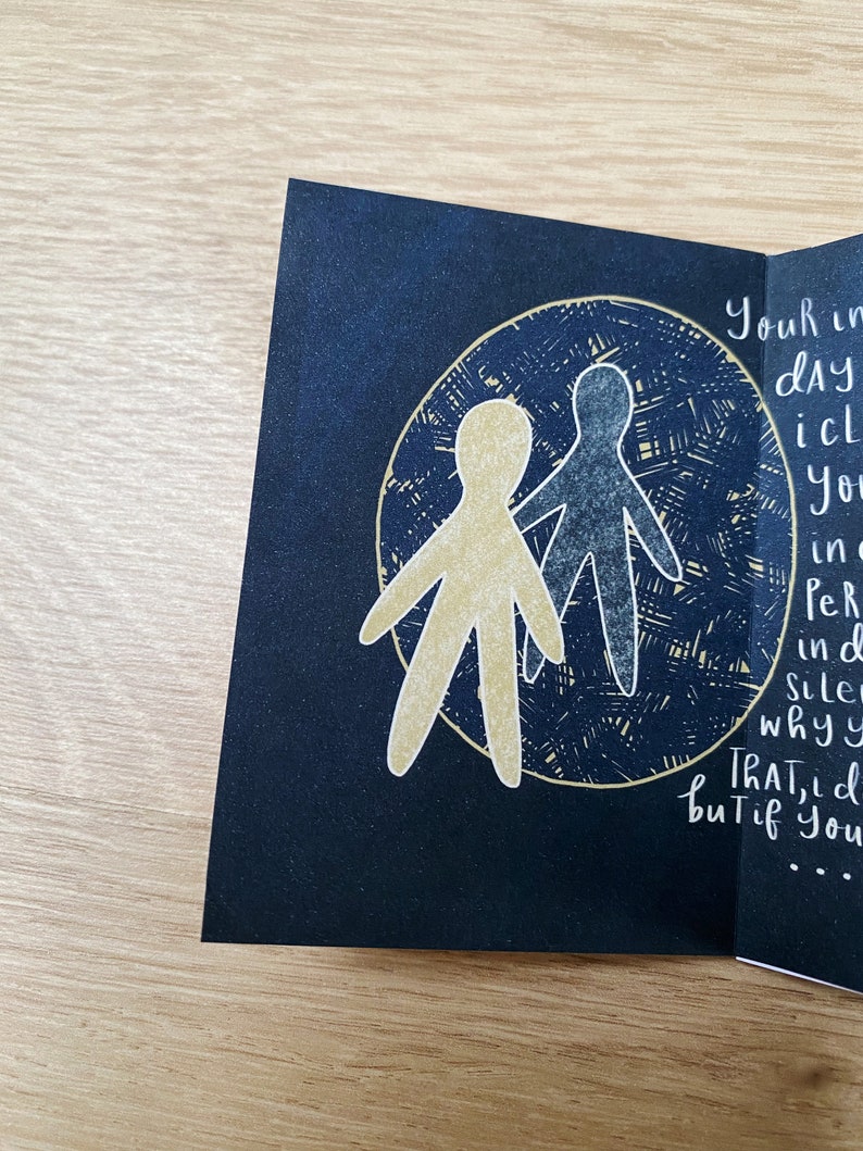 May include: A black card with a gold and blue illustration of two stick figures holding hands inside a circle. The text on the card reads "Your in dAY icl You in Per ind Sile whyy ThAT, d but if You..."