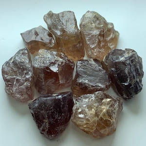 Puede incluir: Un conjunto de nueve piedras preciosas en bruto y sin pulir en tonos variados de marrón translúcido, ámbar y marrón rojizo intenso. Cada piedra presenta una forma irregular y dentada con superficies facetadas que reflejan la luz.