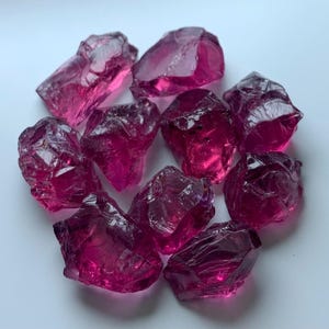 Peut inclure: Un ensemble de pierres précieuses brutes, translucides, de couleur magenta foncé. Les pierres de forme irrégulière sont regroupées, présentant des facettes et des reflets internes. Les gemmes sont posées sur un fond blanc uni.