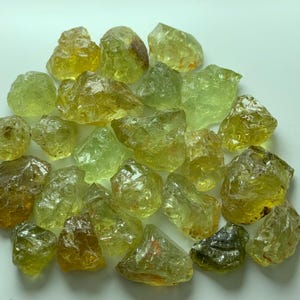 May include: A collection of raw, unpolished gemstones in various shades of yellow, lime green, and olive. These translucent, irregular mineral fragments exhibit natural facets and crystalline textures, displaying a range of vibrant, earthy tones.