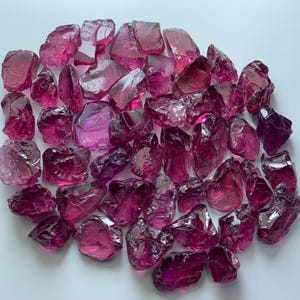 Puede incluir: Una colección de gemas en bruto, sin cortar y de forma irregular, en tonos rosa y magenta. Las gemas están dispersas sobre una superficie blanca, mostrando su apariencia translúcida y cristalina. Las piedras varían en tamaño y forma.