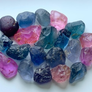 Può includere: Una collezione di pietre preziose grezze in vari colori, tra cui viola scuro, blu, rosa e verde acqua. Le pietre hanno forme irregolari e una consistenza naturale e organica. Le pietre preziose sono disposte vicine, creando un'esposizione visivamente accattivante.
