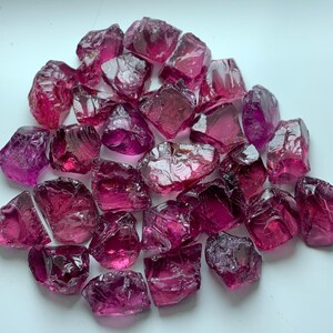 Könnte beinhalten: Eine Ansammlung von rohen, durchscheinenden magentafarbenen Edelsteinfragmenten mit unregelmäßigen, gezackten Formen und facettierten Oberflächen. Diese kleinen, kristallinen Stücke weisen tiefe rosa und violette Farbtöne auf und reflektieren Licht.