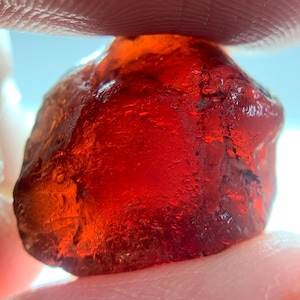 Peut inclure: Une pierre précieuse brute rouge foncé translucide avec une surface irrégulière et à facettes multiples. La pierre présente une teinte cramoisie vibrante et une clarté interne, montrant des textures naturelles et des bords dentelés.