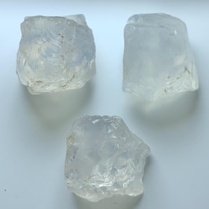 Puede incluir: Tres cristales de cuarzo en bruto, translúcidos y de formas irregulares. Los cristales son de un color blanco lechoso pálido y parecen estar tallados en bruto. La imagen está tomada desde arriba, con los cristales dispuestos sobre una superficie blanca.