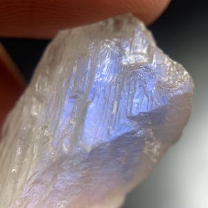 May include: Close-up of a raw, translucent moonstone crystal. The stone has a rough, textured surface with visible striations. The moonstone displays a subtle play of light, with flashes of blue and iridescent hues.
