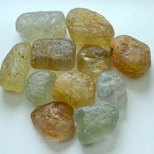 May include: A collection of ten raw, unpolished gemstones in various shapes and sizes. The stones display a range of translucent and opaque colours, including pale blue, light yellow, golden amber, and soft green, with natural textured surfaces.