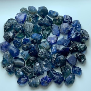 Può includere: Una collezione di zaffiri grezzi e non lucidati in varie tonalità di blu profondo, blu navy e verde acqua scuro. Le pietre sono di forma irregolare con superfici sfaccettate e strutturate, che mostrano un mix di finiture traslucide e opache.