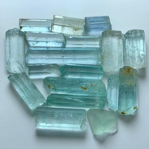 Puede incluir: Un conjunto de cristales de aguamarina en bruto, de diversas formas y tamaños. Los cristales varían en color, desde azul pálido hasta turquesa claro, con inclusiones naturales. Las piedras preciosas están dispuestas sobre una superficie blanca.