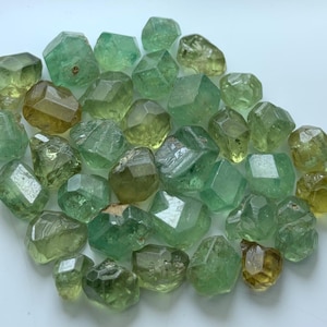 Pode incluir: Uma coleção de pedras preciosas facetadas soltas em vários tons de verde e verde-amarelado. Estas pequenas pedras de múltiplas facetas exibem uma qualidade translúcida com superfícies polidas, apresentando formas geométricas irregulares e variações naturais de cor.