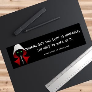 Puede incluir: Pegatina negra y roja con una silueta blanca de una mujer que lleva una capa roja y una capucha blanca. El texto de la pegatina dice "IGNORING ISN'T THE SAME AS IGNORANCE, YOU HAVE TO WORK AT IT. ATWOOD, M. (1985). THE HANDMAID'S TALE."