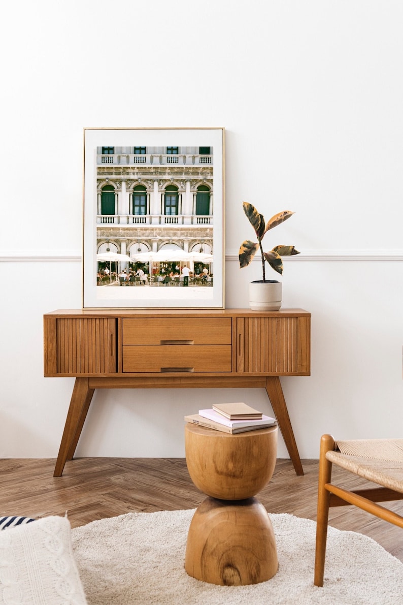 Op de afbeelding: Een ingelijste foto van een gebouw met groene ramen en witte luifels. Het gebouw bevindt zich in een stad en er zitten mensen aan tafels buiten. De foto staat op een houten dressoir met een plant in een pot en een houten kruk met boeken erop.