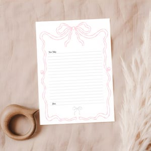 Może przedstawiać: Biała karta papieru listowego z r&oacute;żowym wzorem kokardy i liniowaną przestrzenią do pisania. Tekst "Dear Baby" znajduje się na g&oacute;rze, a "Love" na dole. Drewniany gryzak znajduje się w lewym dolnym rogu.