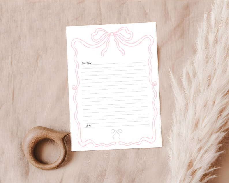 Może przedstawiać: Biała karta papieru listowego z r&oacute;żowym wzorem kokardy i liniowaną przestrzenią do pisania. Tekst "Dear Baby" znajduje się na g&oacute;rze, a "Love" na dole. Drewniany gryzak znajduje się w lewym dolnym rogu.