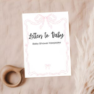 Może przedstawiać: Biała karta z napisem "Letters to Baby Baby Shower Keepsake" w czarnym skrypcie, obramowana r&oacute;żowym wzorem kokardy. Drewniana grzechotka dla dziecka i puszysta, suszona roślina r&oacute;wnież znajdują się na zdjęciu.