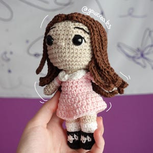 May include: Handmade crochet doll with brown hair and large black eyes. The doll wears a pink dress with a white collar and black shoes. The doll is held in a hand against a purple background.