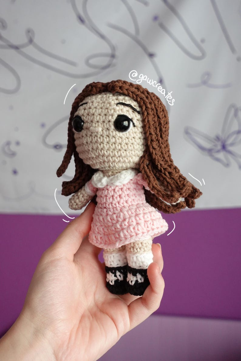 May include: Handmade crochet doll with brown hair and large black eyes. The doll wears a pink dress with a white collar and black shoes. The doll is held in a hand against a purple background.