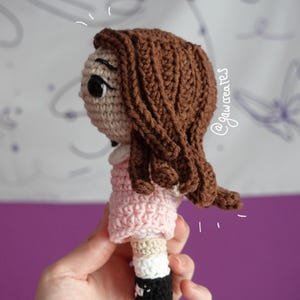 May include: Crocheted doll with brown hair, a light pink dress, and black and white shoes. The doll's face is beige with black eyes. The doll is handmade and appears to be a child's toy.