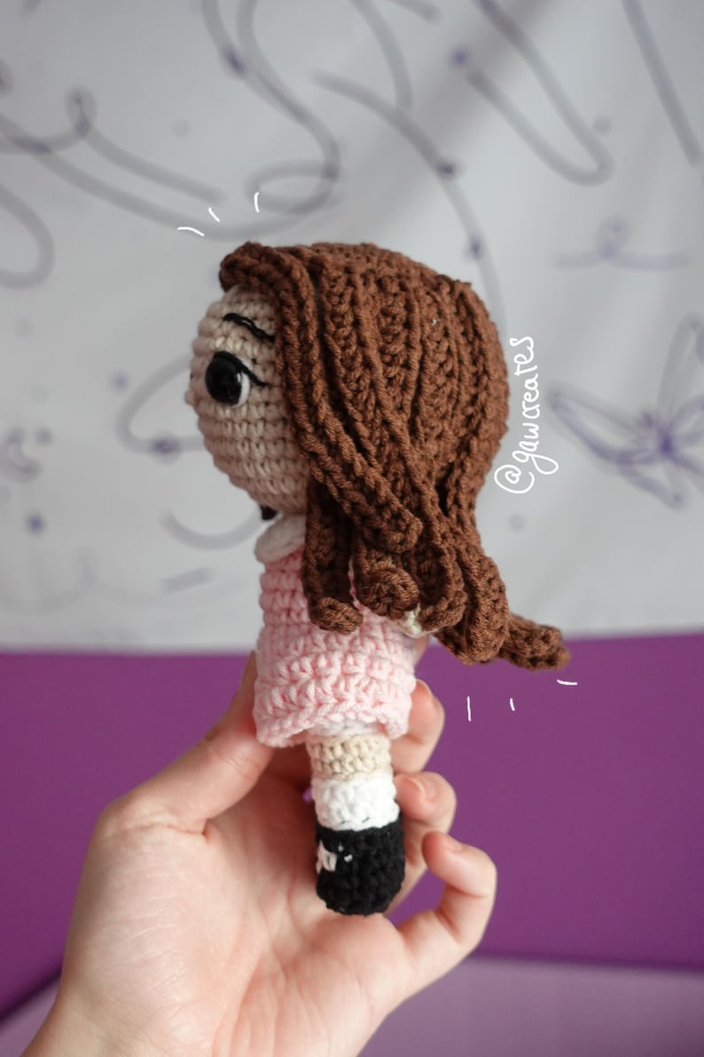 May include: Crocheted doll with brown hair, a light pink dress, and black and white shoes. The doll's face is beige with black eyes. The doll is handmade and appears to be a child's toy.