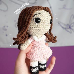 May include: A handmade crochet doll with brown hair and large black eyes. The doll wears a light pink dress with a white collar and black and white shoes. The doll is a charming, handcrafted item.