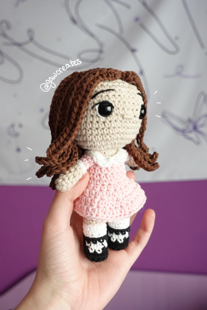 May include: A handmade crochet doll with brown hair and large black eyes. The doll wears a light pink dress with a white collar and black and white shoes. The doll is a charming, handcrafted item.