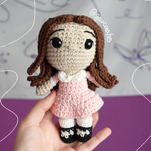 May include: A handmade crochet doll with brown hair and large black eyes. The doll wears a pink dress with a white collar, white socks, and black shoes. The doll is held in a hand. The text "@gawcreates" is visible.