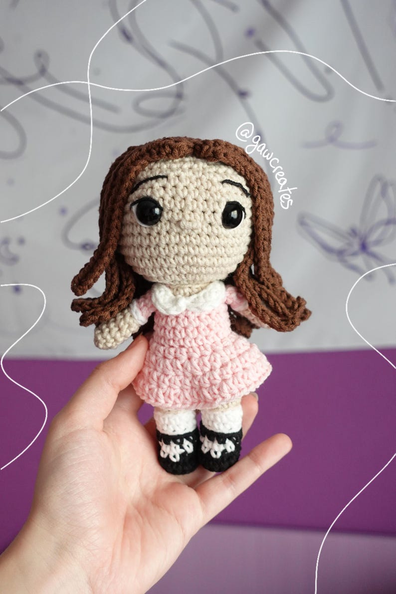 May include: A handmade crochet doll with brown hair and large black eyes. The doll wears a pink dress with a white collar, white socks, and black shoes. The doll is held in a hand. The text "@gawcreates" is visible.