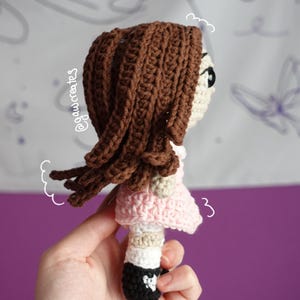 May include: Crocheted doll with brown hair, a pink dress, and black shoes. The doll has a cream-colored face and is held in a hand. The doll's hair is styled with long, flowing strands. The doll is a handmade item.