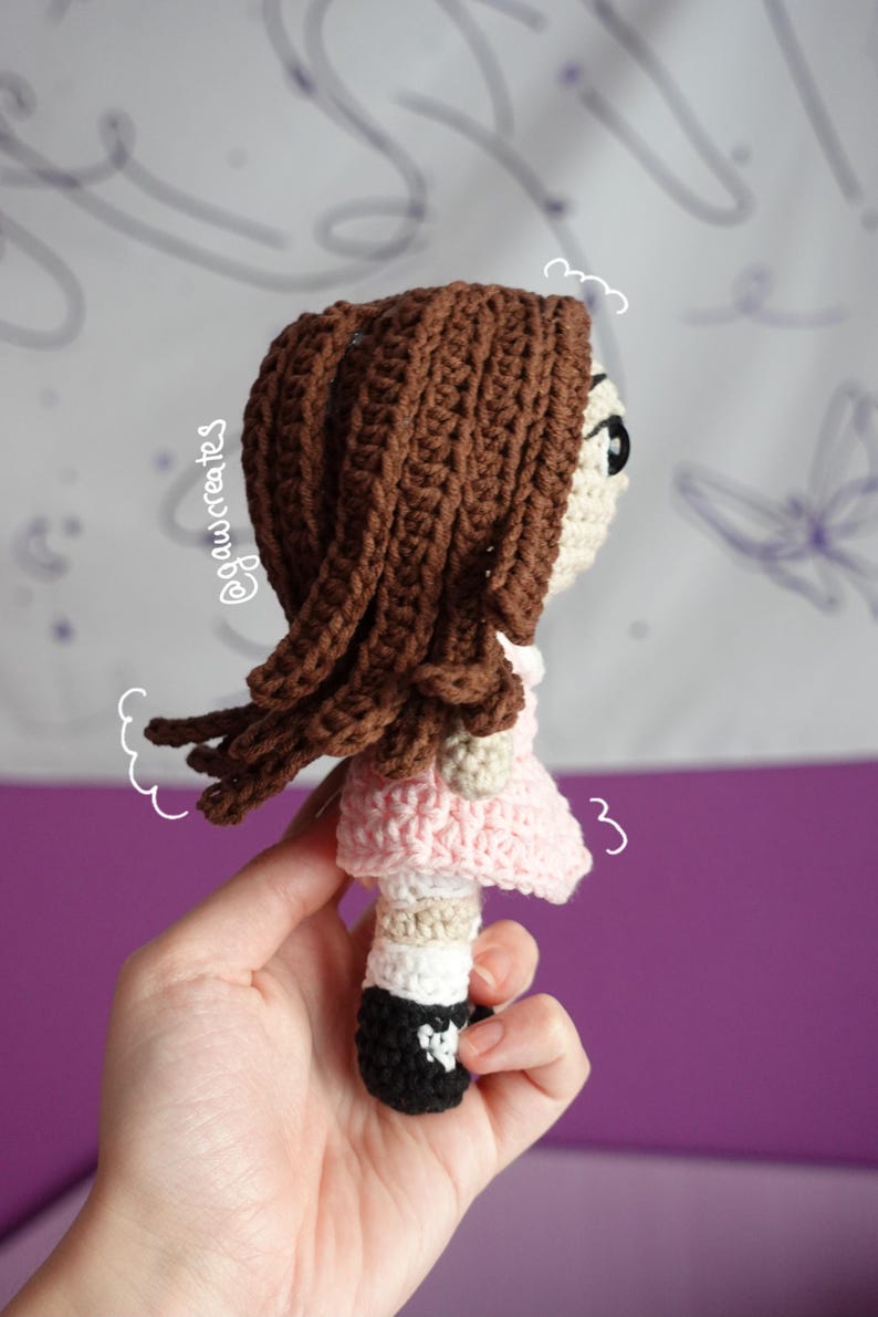 May include: Crocheted doll with brown hair, a pink dress, and black shoes. The doll has a cream-colored face and is held in a hand. The doll's hair is styled with long, flowing strands. The doll is a handmade item.