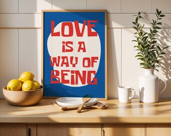 Puede incluir: Una impresión artística enmarcada con las palabras "LOVE IS A WAY OF BEING" en letras rojas sobre un fondo azul. Un cuenco de madera con limones, platos blancos y un jarrón blanco con vegetación están sobre una encimera de madera.
