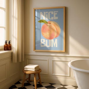 Op de afbeelding: Een ingelijste kunstprint met een lichtblauwe achtergrond en de woorden "NICE BUM" in witte blokletters. Het kunstwerk toont een perzik met een groen blad. De lijst is van lichtgekleurd hout.