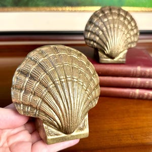 May include: A pair of brass bookends shaped like scallop shells. Each bookend features a textured, fan-like design with radiating ridges and a solid, rectangular base. The metal has a warm, metallic gold finish with visible detailing.