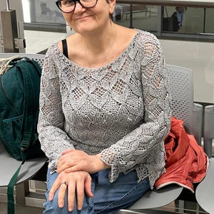 May include: A gray, open-weave sweater with a floral pattern. The long-sleeved top is paired with blue jeans. The person is wearing black-framed glasses and a necklace. A green backpack and red jacket are visible.