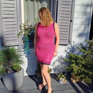 May include: A sleeveless, knee-length dress in a vibrant hot pink hue, featuring an intricate, textured knit pattern throughout. The garment is accented with a contrasting black trim along the neckline, armholes, and hem, adorned with small black beads.