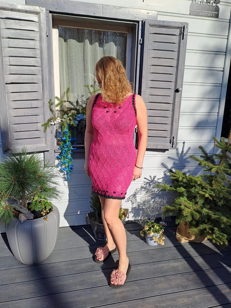 May include: A sleeveless, knee-length dress in a vibrant hot pink hue, featuring an intricate, textured knit pattern throughout. The garment is accented with a contrasting black trim along the neckline, armholes, and hem, adorned with small black beads.