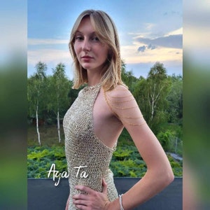 May include: A sleeveless, high-neck dress crafted from a metallic gold, open-knit mesh fabric. The garment features a delicate gold-toned multi-strand chain shoulder accessory that drapes across the upper arm. The text "Aga Ta" is visible in white.