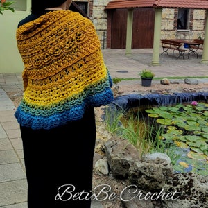 Puede incluir: Un chal tejido a crochet con un degradado de colores amarillo, verde y azul. El chal está drapeado sobre los hombros de una persona y está hecho con un delicado patrón de puntadas. El chal se muestra frente a un estanque con nenúfares.