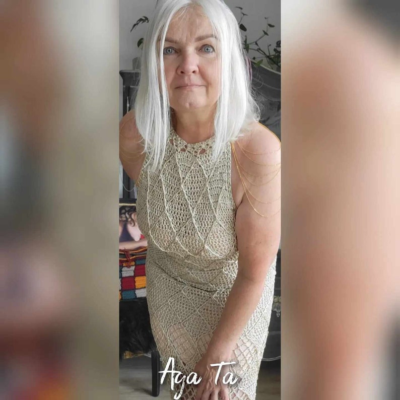 May include: A beige sleeveless crochet dress featuring a diamond-shaped pattern throughout the bodice and skirt. The garment has a high neckline and is accented with delicate gold-toned chain straps draped over the shoulders. The text Aga Ta is visible.