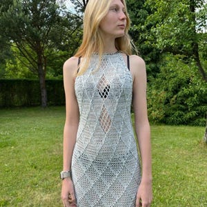 May include: A sleeveless, floor-length crochet dress in a light silver-grey hue. The garment features a repeating diamond-shaped openwork pattern throughout the body, a high halter-style neckline, and a fitted silhouette that reaches the ankles.
