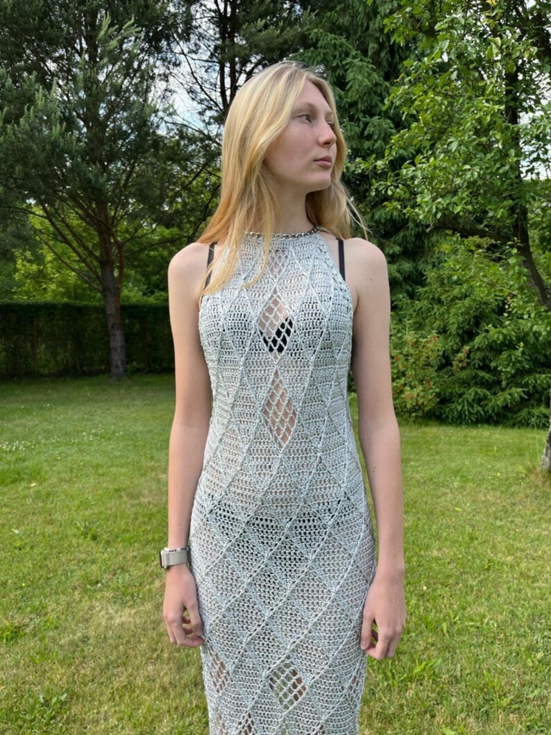 May include: A sleeveless, floor-length crochet dress in a light silver-grey hue. The garment features a repeating diamond-shaped openwork pattern throughout the body, a high halter-style neckline, and a fitted silhouette that reaches the ankles.