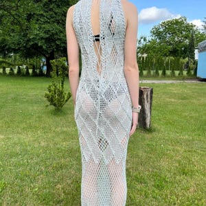 May include: A floor-length, sleeveless crochet dress in a light silver-grey hue. The garment features an intricate, open-work diamond pattern throughout, a high neckline, and a vertical lace-up detail along the back that exposes the skin.