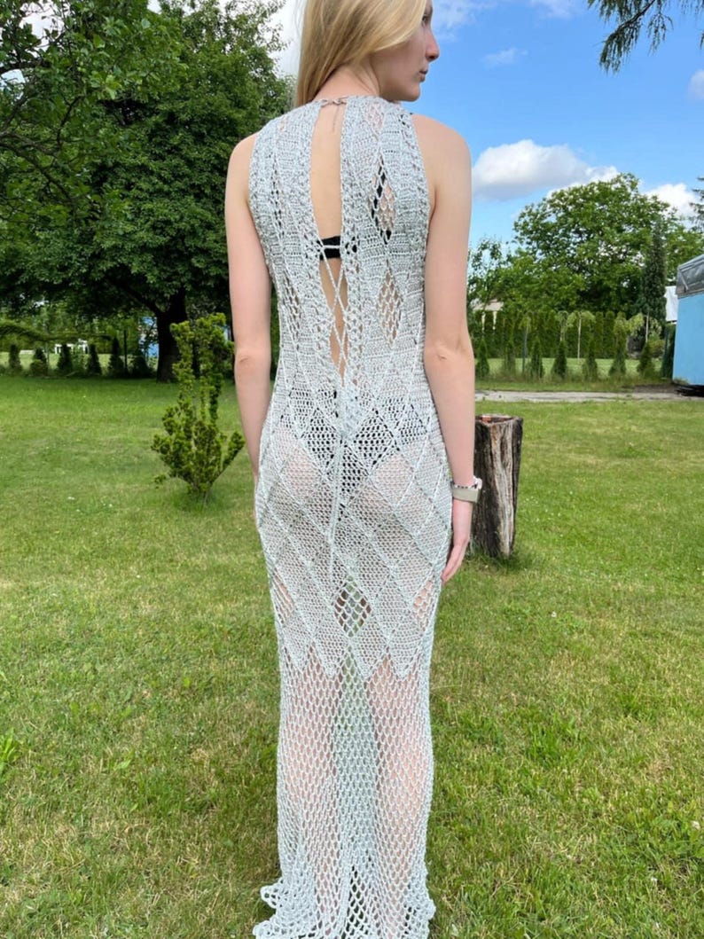 May include: A floor-length, sleeveless crochet dress in a light silver-grey hue. The garment features an intricate, open-work diamond pattern throughout, a high neckline, and a vertical lace-up detail along the back that exposes the skin.