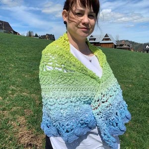 May include: A crocheted shawl in shades of green, blue, and white. The shawl is draped over the shoulders, with a scalloped edge. The background features a green field, blue sky, and houses.